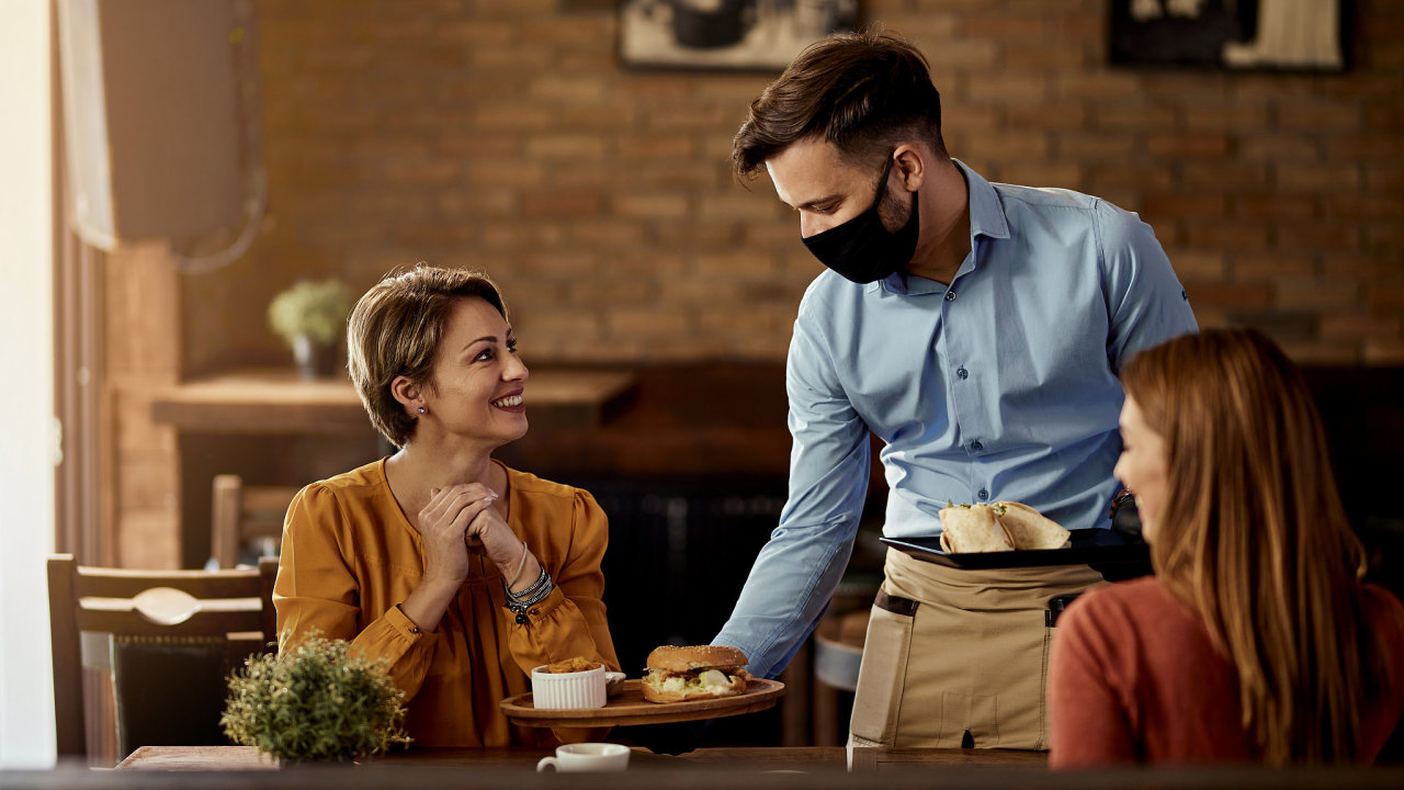 Young waiter wearing protective face mask while serving food to his guests in a restaurant.
