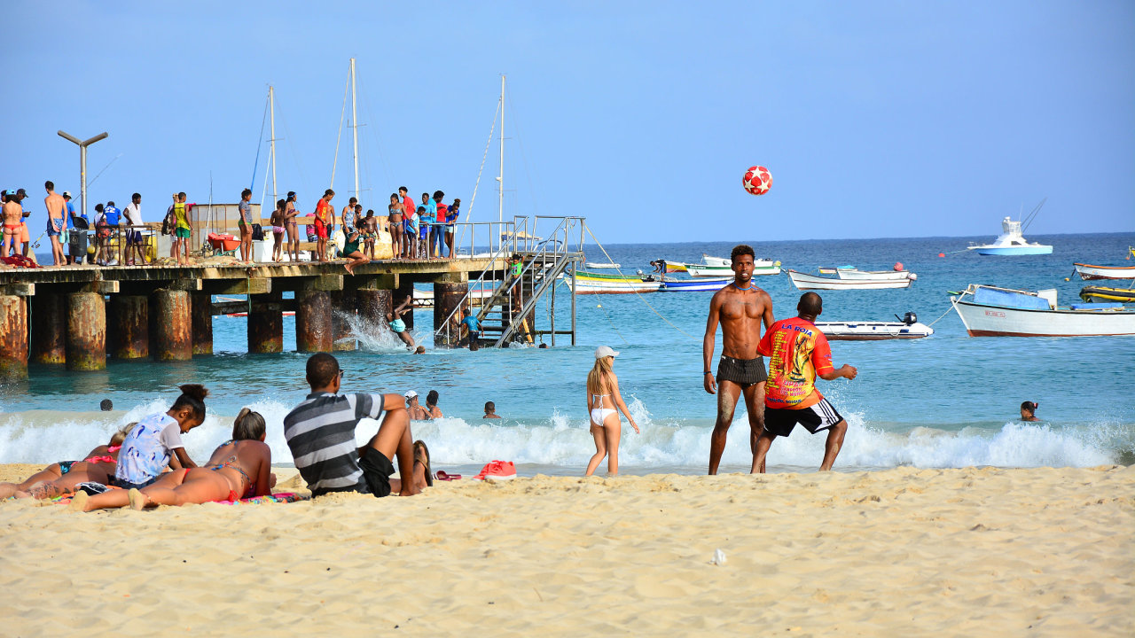 Santa Maria, Sal, Cape Verde, Cabo Verde, July 10th, 2019 - People relaxing and playing football on sandy beach, wooden pier and colorful traditional fishing boats